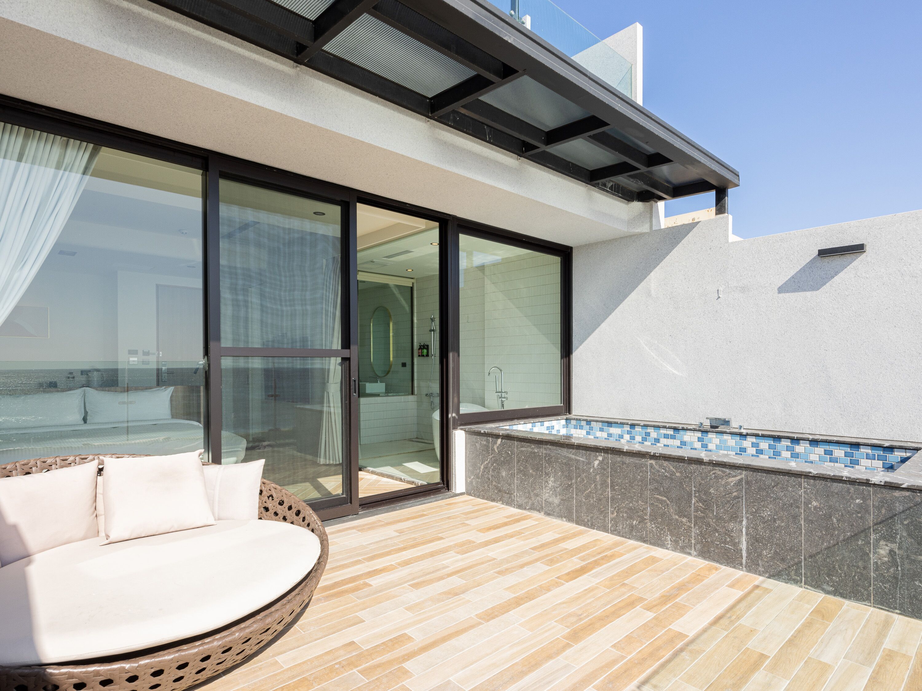 Un espacio exterior moderno con una tumbona, un ventanal de vidrio que da a un baño y una pequeña piscina.