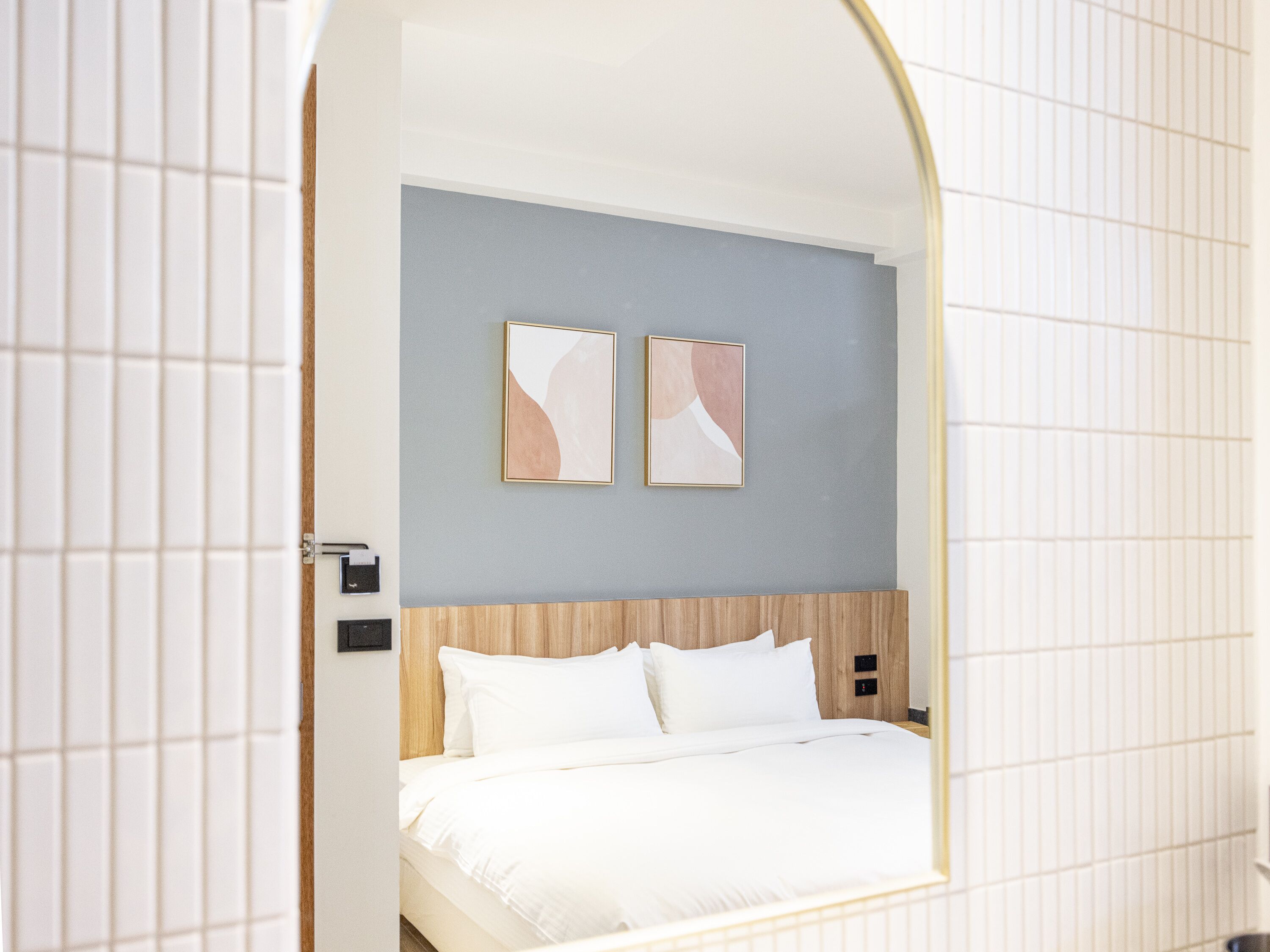Habitación de hotel con cama, cabecera de madera, dos cuadros abstractos colgados en la pared y pared de azulejos blancos.