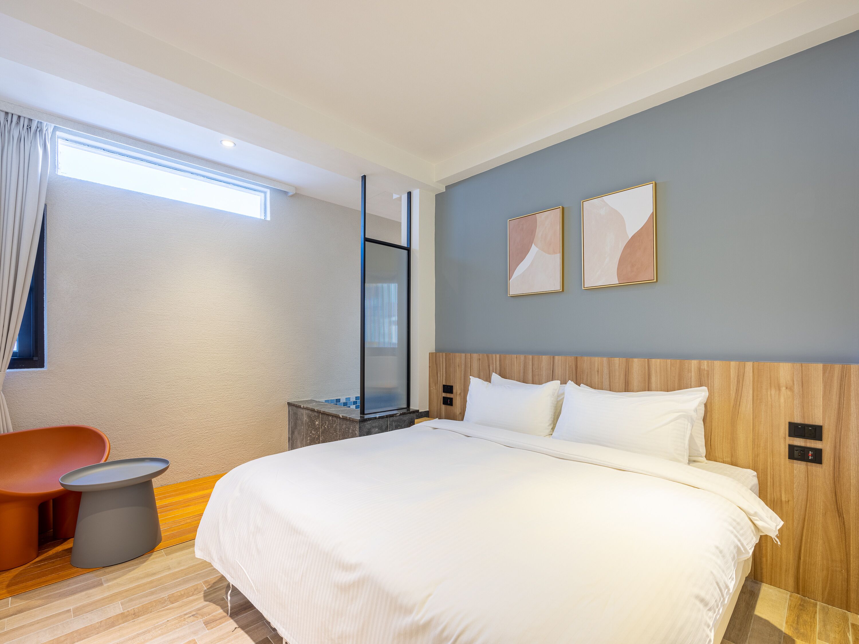 A modern hotel room with a large bed, a wooden headboard, a small side table, and a window with curtains.