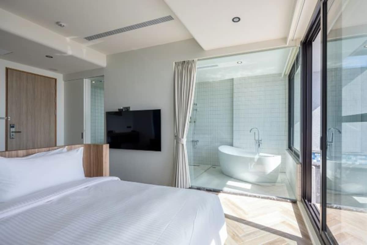 A modern hotel room with a large bed, a TV mounted on the wall, a glass shower enclosure, and a view of the city.