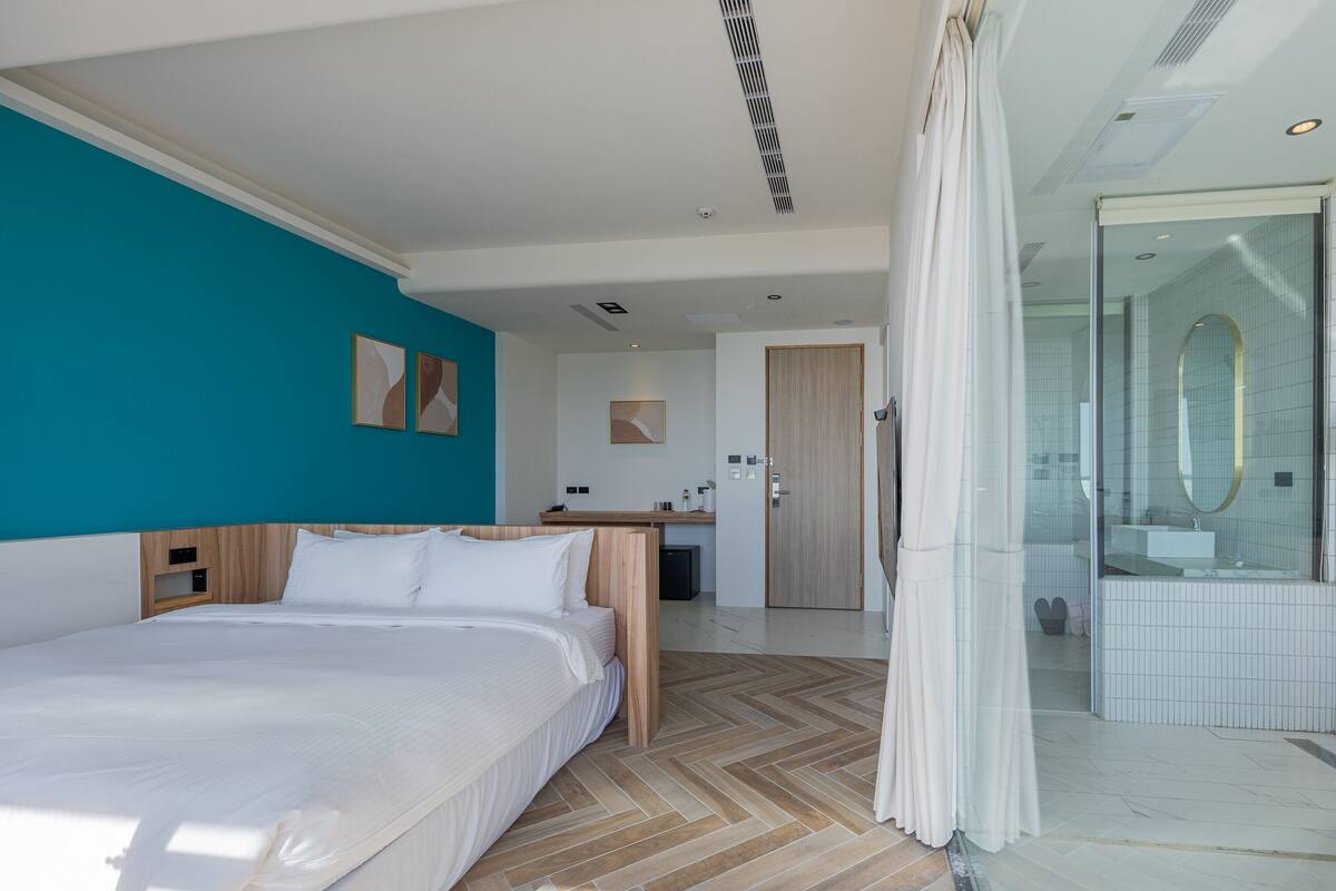 Ett modernt hotellrum med en stor säng, en sänggavel i trä, en vägg i en turkos nyans som accentfärg, ett badrum med en dusch av glas och ett golv med fiskbensmönster.