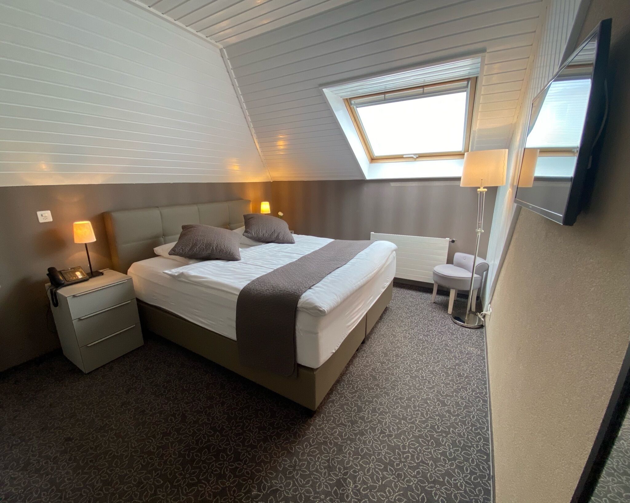 Une chambre d’hôtel avec un grand lit, des lampes de chevet, une petite chaise, un bureau avec un téléphone et une fenêtre avec des stores.
