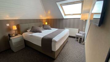 Une chambre d’hôtel avec un grand lit, des lampes de chevet, une petite chaise, un bureau avec un téléphone et une fenêtre avec des stores.