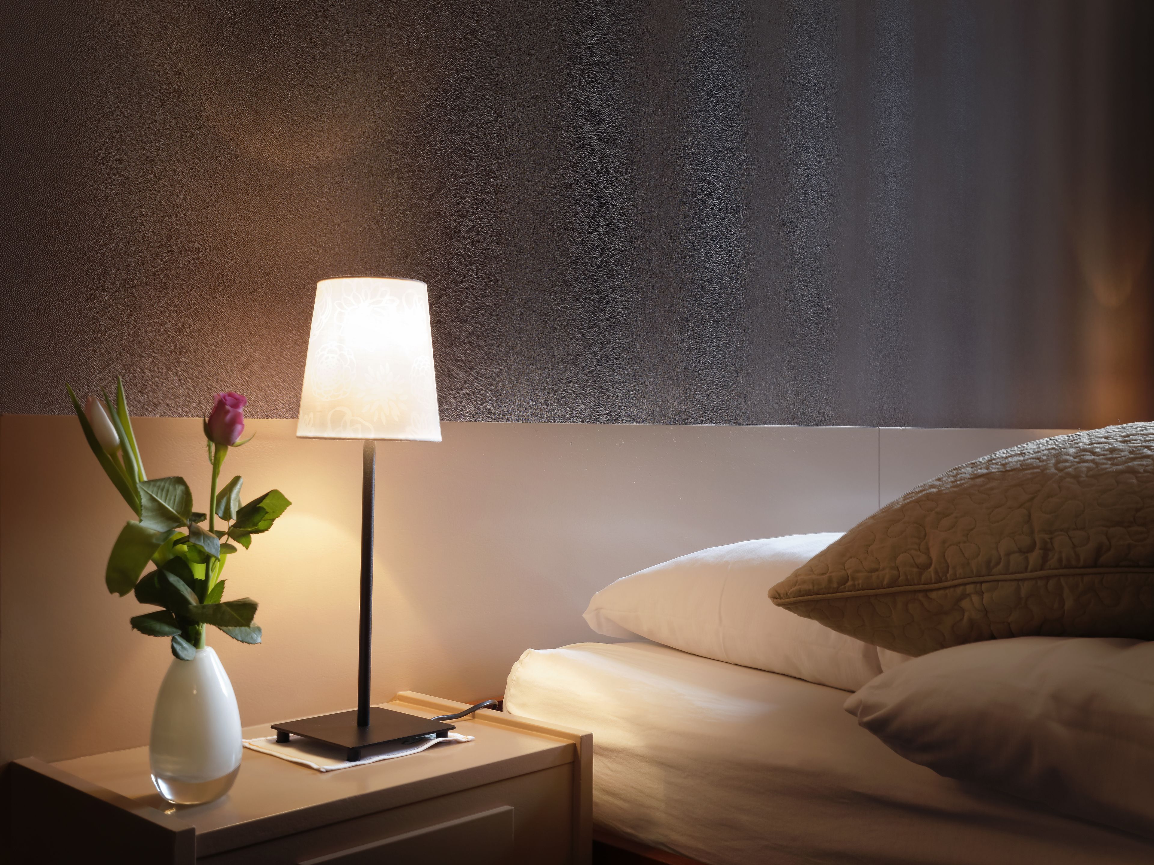Una cama bien hecha con un almohadón blanco y otro color crema acolchado. Una mesita de noche sostiene un florero blanco con una rosa y una lámpara con pantalla blanca.