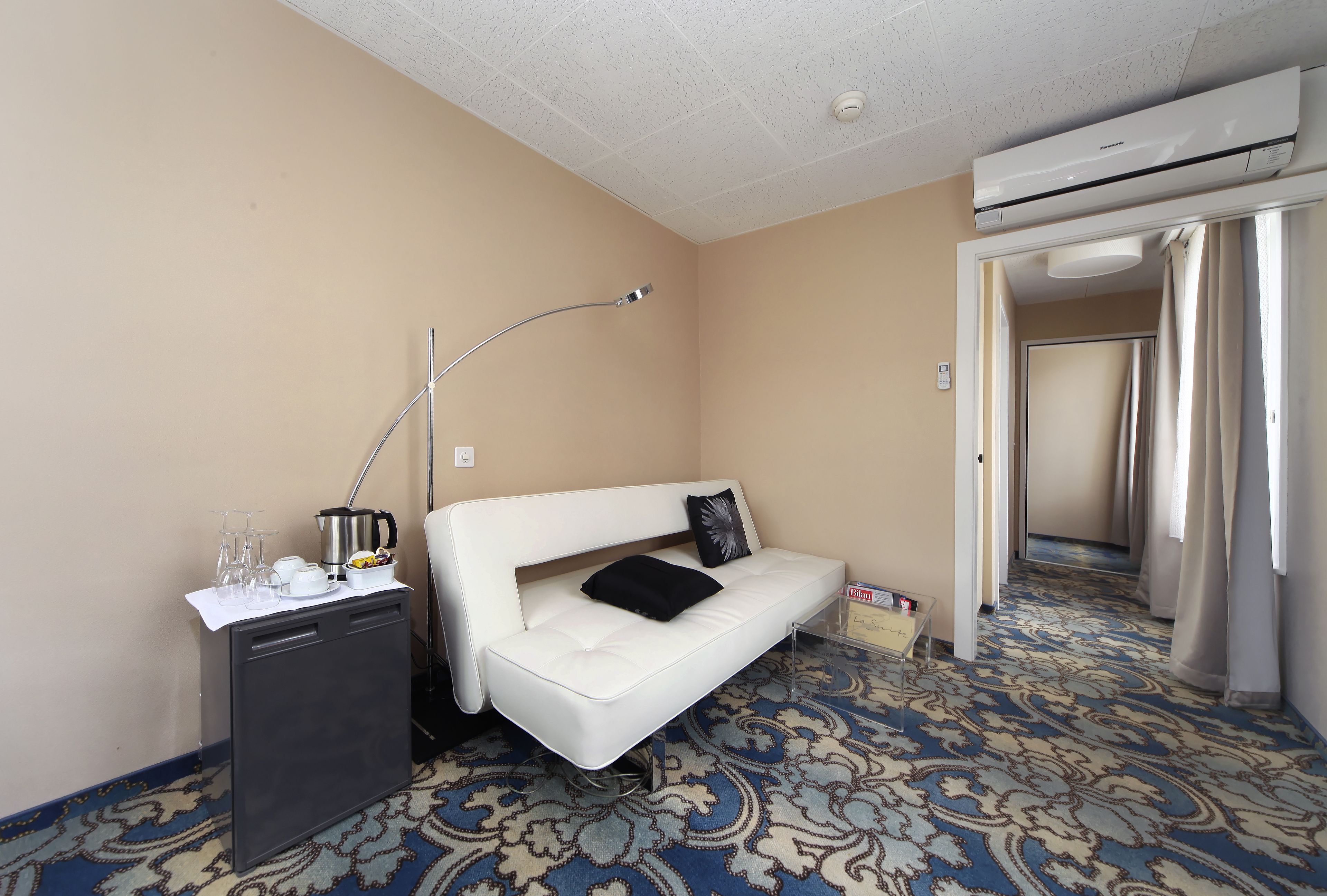 Une chambre d’hôtel avec un canapé blanc, une petite table avec une lampe, un minibar et une moquette à motifs.