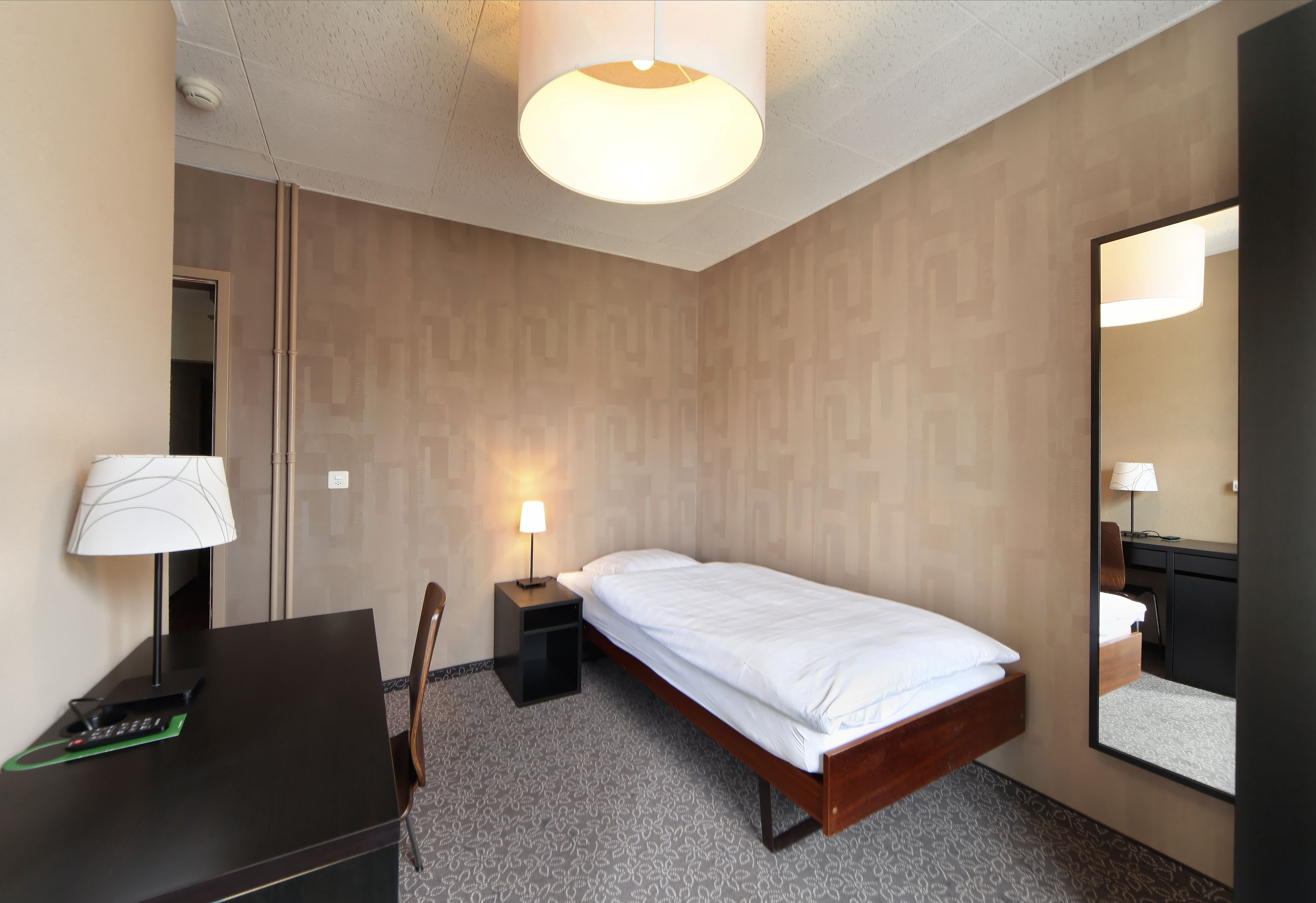 A hotel room with a bed, bedside table, lamp, desk, chair, and a large mirror.