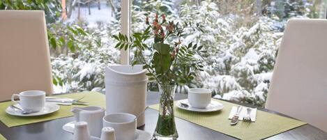 A dining table set with white cups, saucers, and a pitcher, with a vase of greenery in the center.