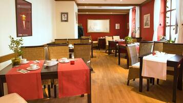 Un'area pranzo con tavoli in legno e sedie in vimini, tovaglie rosse e opere d'arte incorniciate alle pareti.