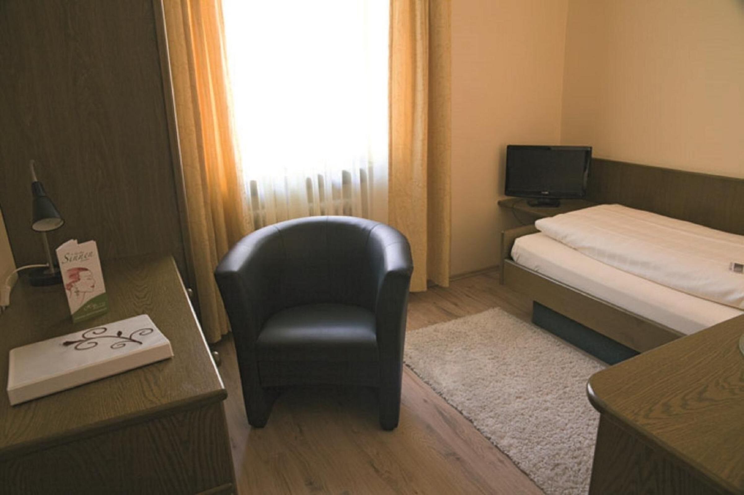 A hotel room with a wooden desk, a black leather chair, a bed, a television, and a window with curtains.