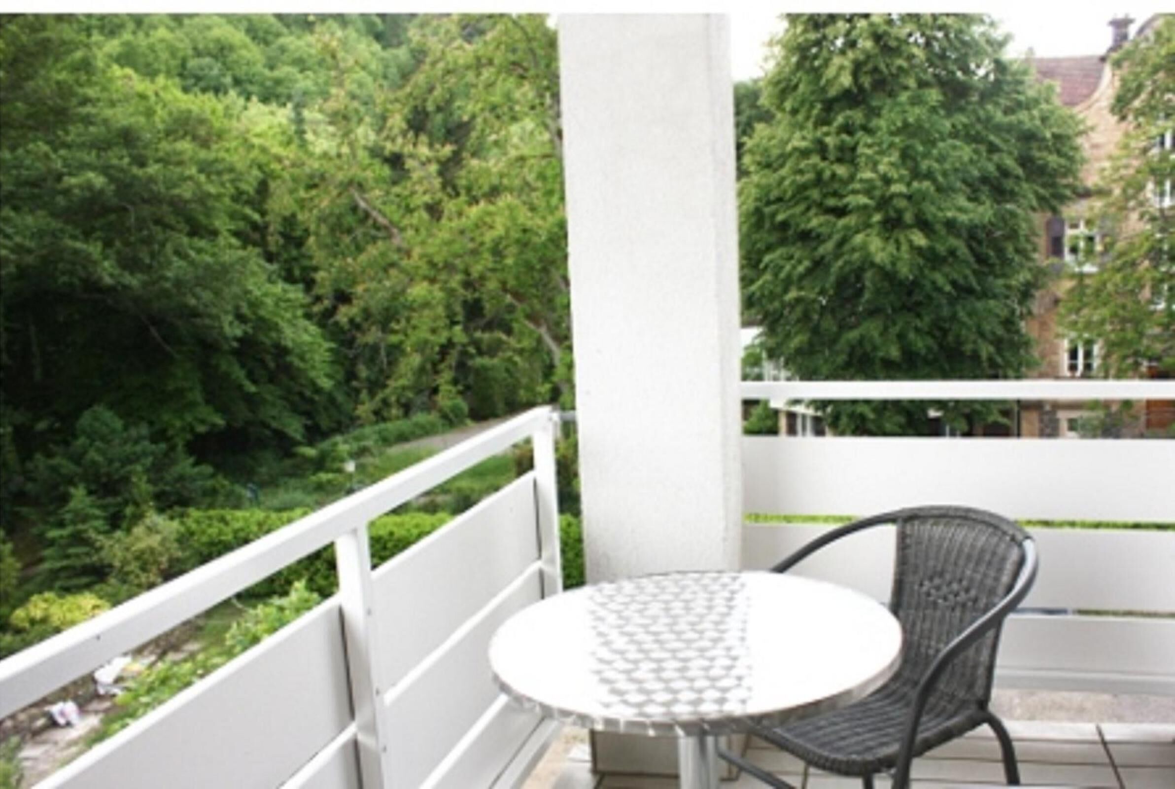 A balcony with a round table and a wicker chair, surrounded by greenery and buildings.