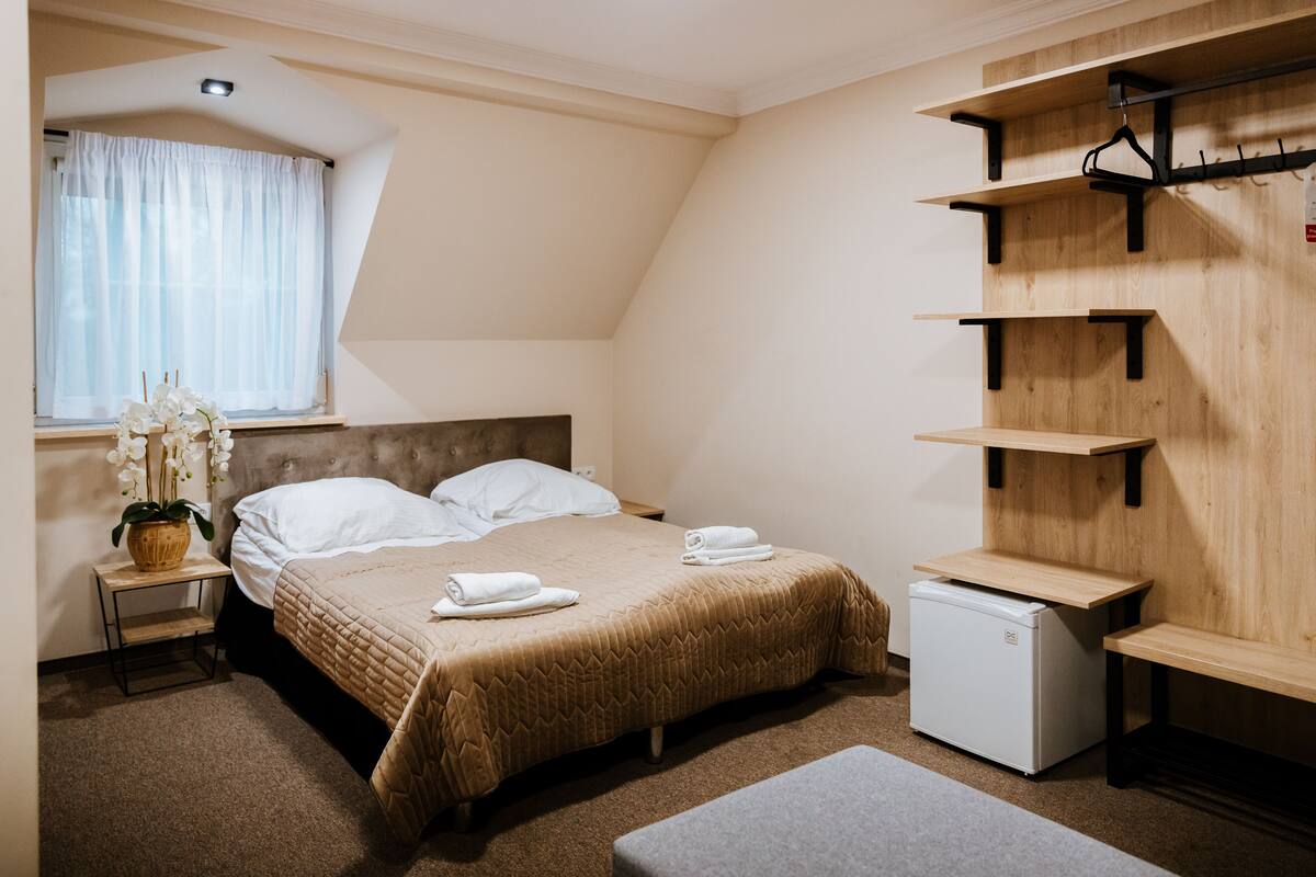 A bedroom with a bed, a small refrigerator, a bedside table with a plant, and a window with curtains.