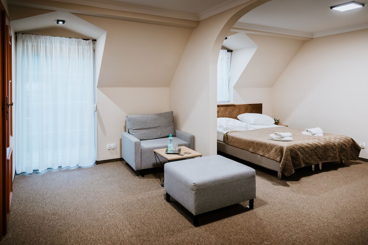 A hotel room with a bed, a grey armchair, a small table with a bottle, and a grey ottoman.