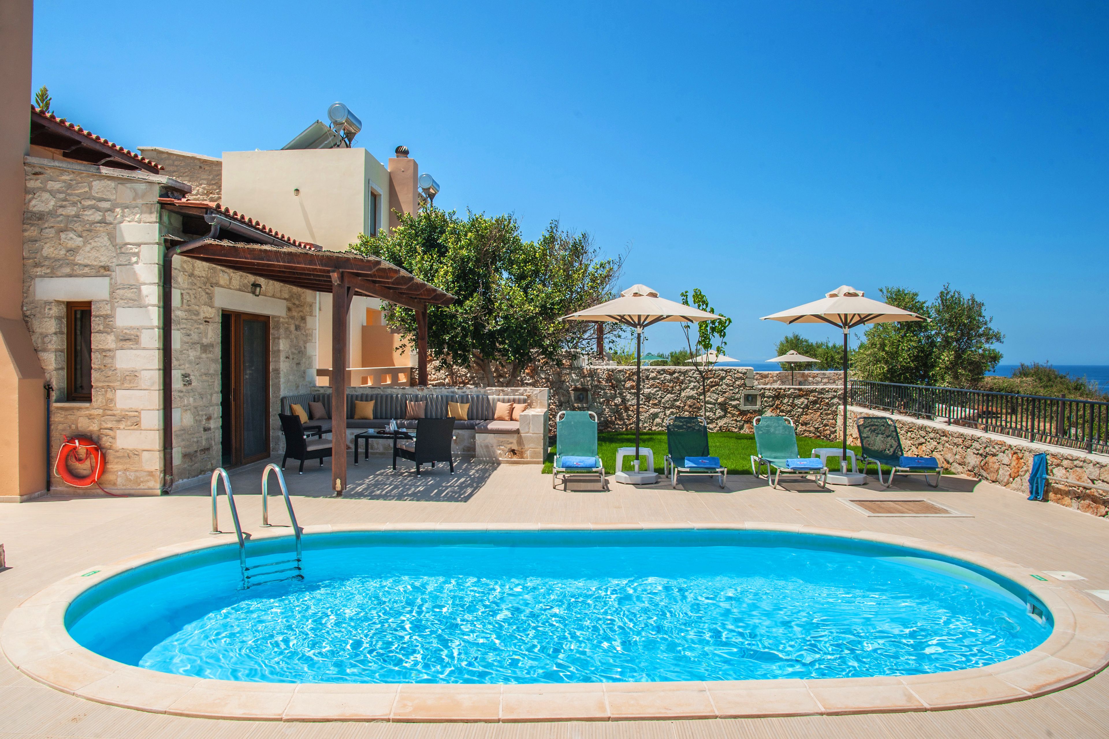 A swimming pool with lounge chairs and umbrellas, surrounded by stone buildings and a clear sky.