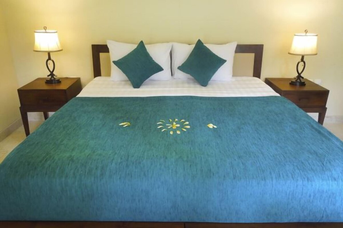Una cama bien tendida con una colcha azul verdosa, dos almohadas verdes y un estampado floral decorativo en la colcha.