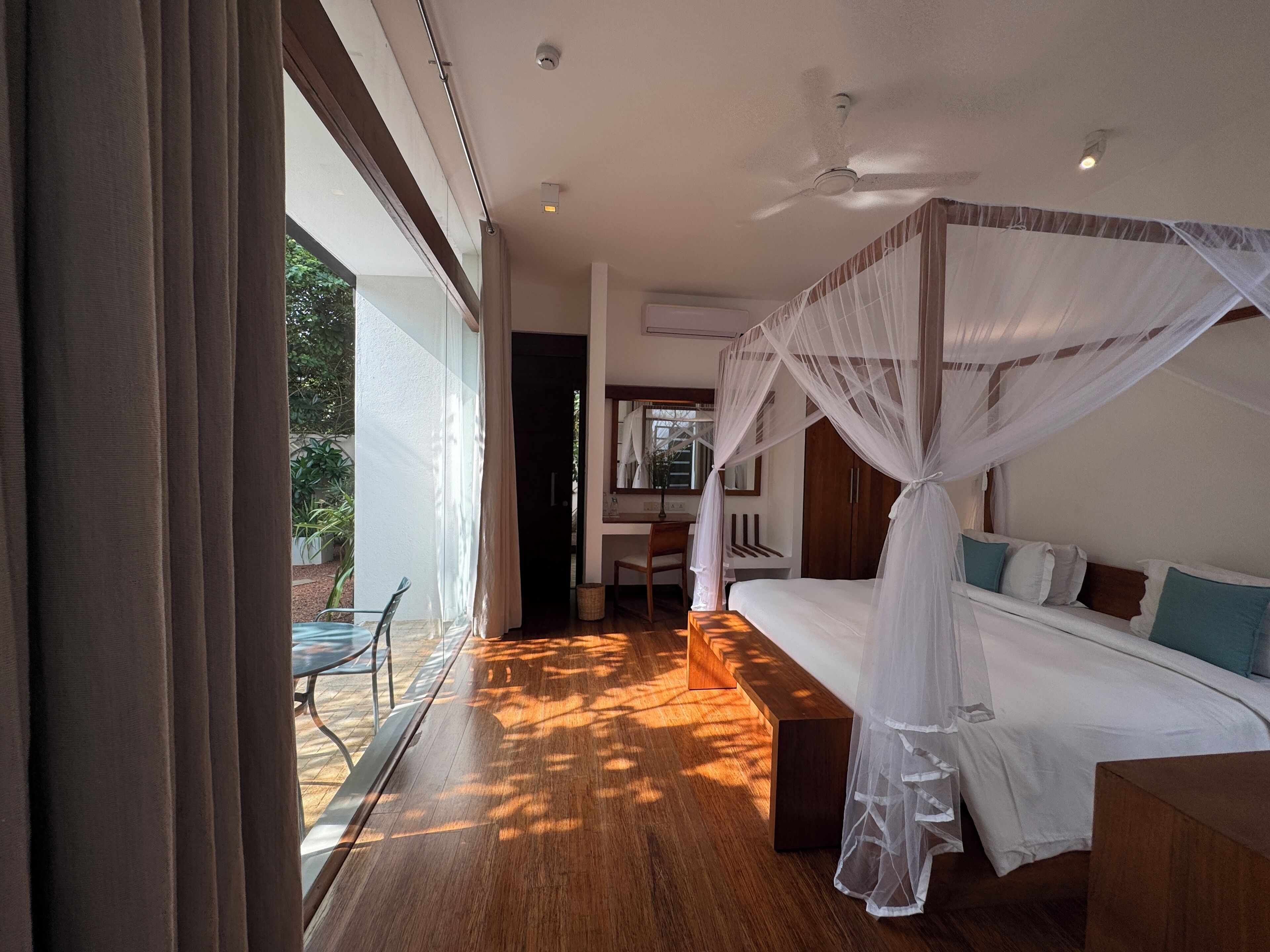 A bedroom with a canopy bed, wooden flooring, a desk, and a view of the outdoors.