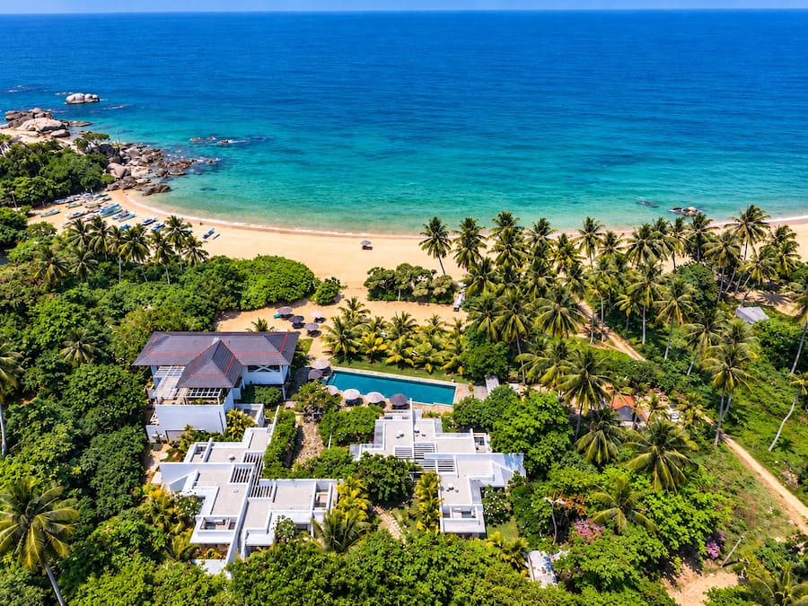 Un resort con varias edificaciones, una piscina y una playa con palmeras.