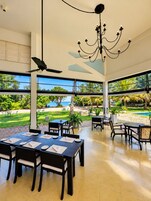 A spacious indoor area with a dining setup, large windows offering a view of a pool and greenery, and a chandelier.