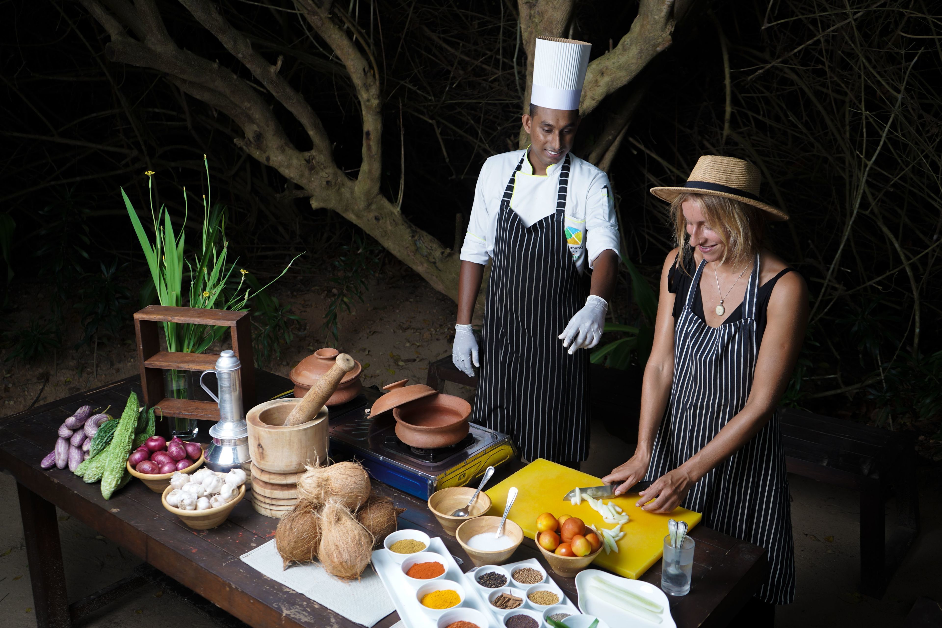 A table with various cooking ingredients and utensils, a person slicing vegetables, and another person standing nearby.