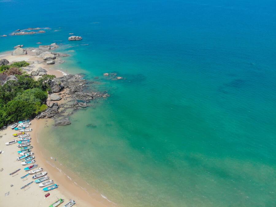 Vue aérienne d’une plage aux eaux turquoise et limpides, avec des formations rocheuses et plusieurs bateaux alignés sur le rivage.