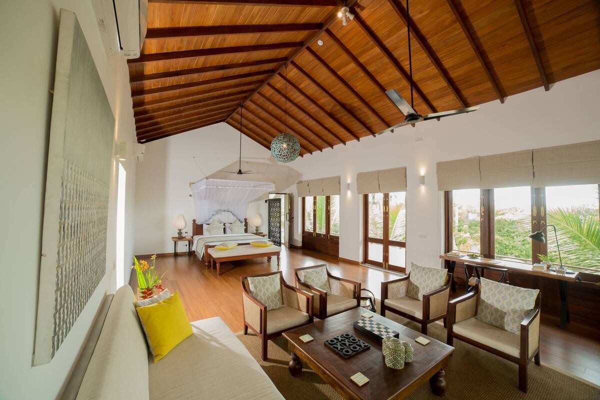 A spacious room with a high ceiling, a bed with a canopy, a dining area with chairs, and large windows offering a view.