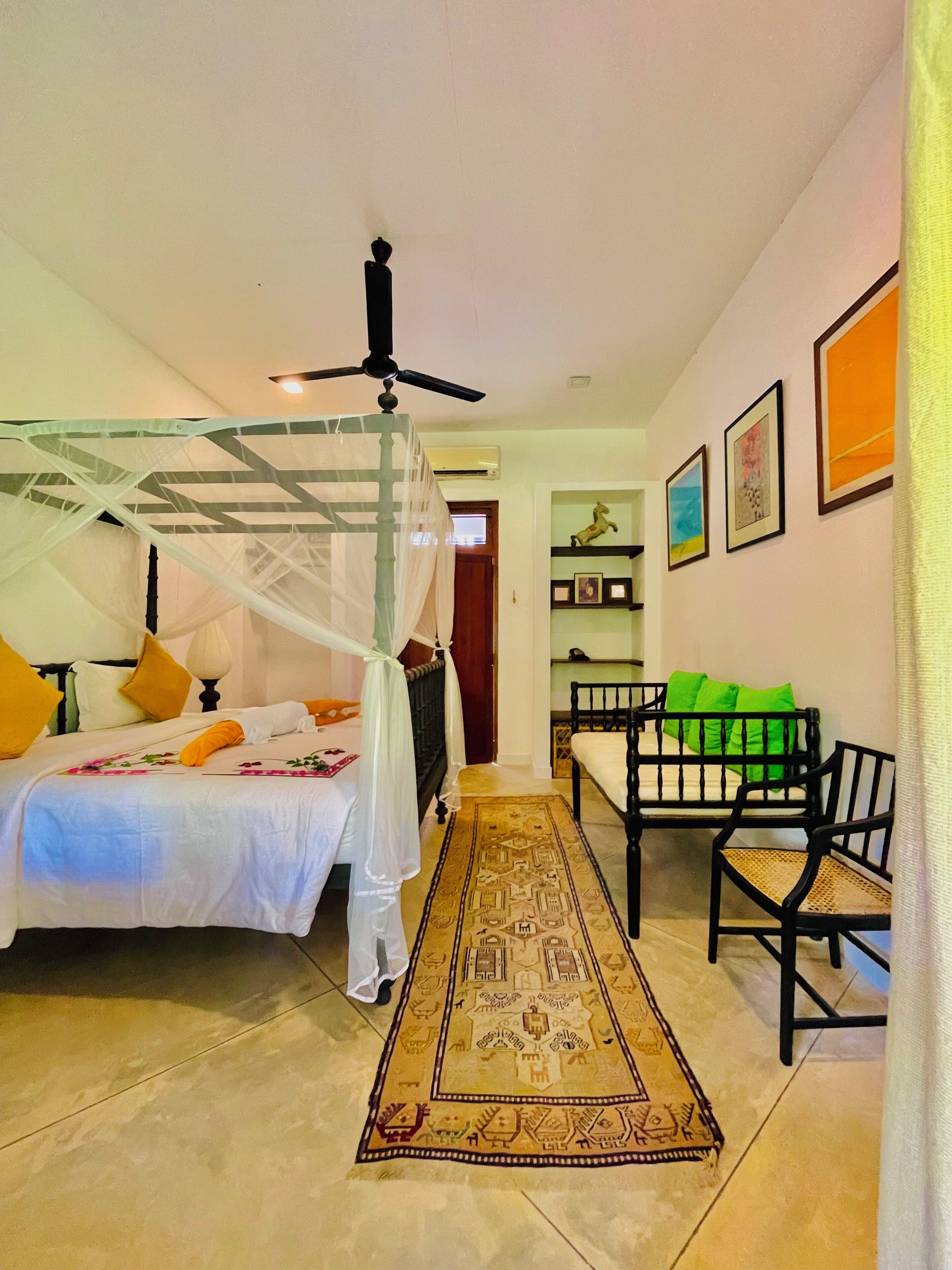 A bedroom with a bed, a daybed, a standing chair, a ceiling fan, and a patterned rug.
