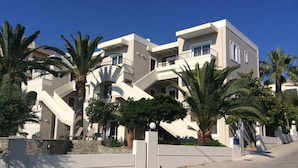 Un immeuble moderne à plusieurs étages, doté de balcons, de palmiers et surplombant un ciel d’un bleu limpide.