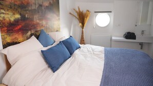 A bedroom with a large bed, white and blue bedding, a painting on the wall, a tall vase with dried plants, and a round window. - Hotel Restaurant Brasserie Ahoi, Kortrijk (Kortrijk)
