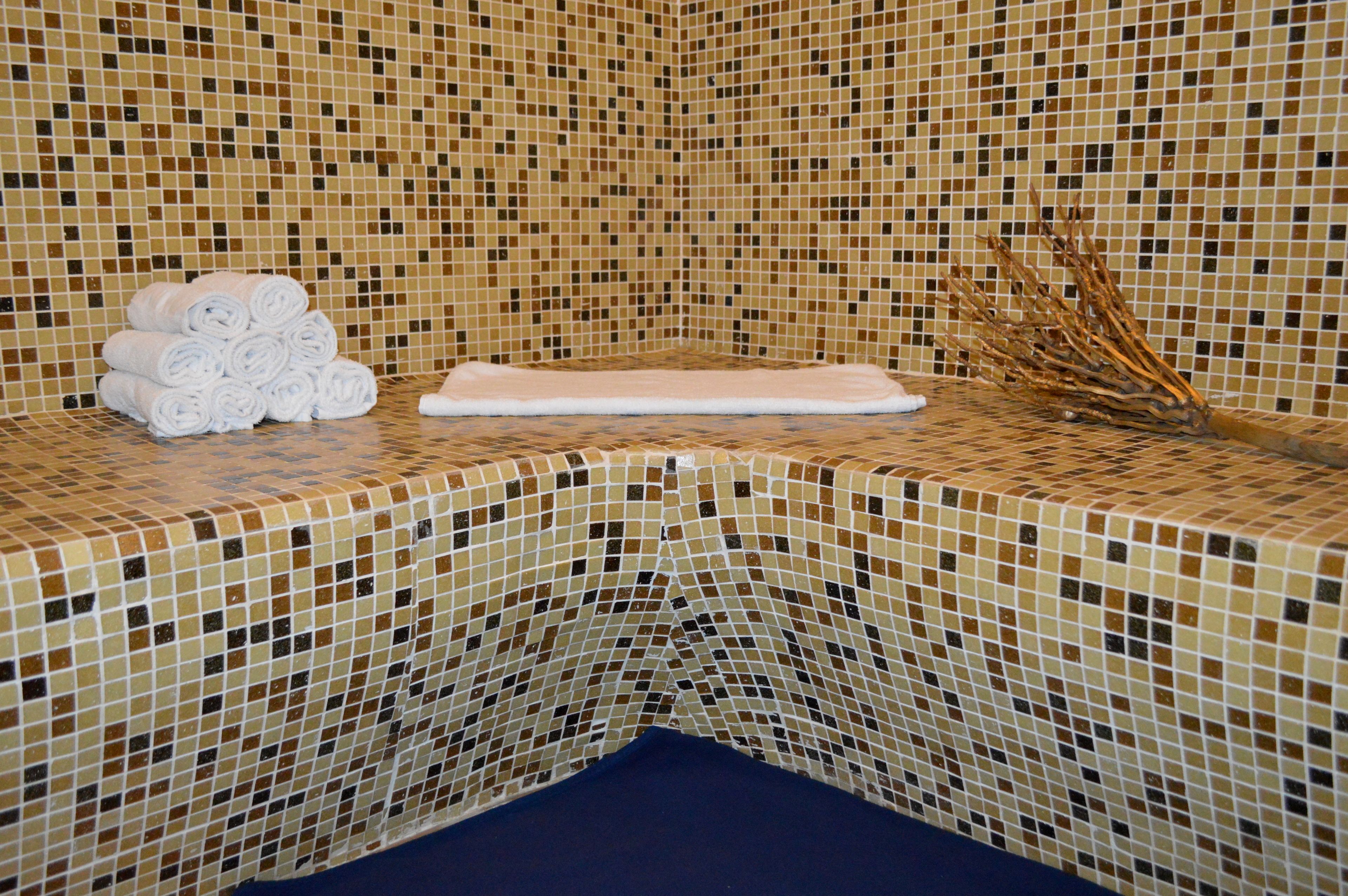Un coin de pièce avec un mur recouvert de mosaïques, une pile de serviettes et une branche décorative.