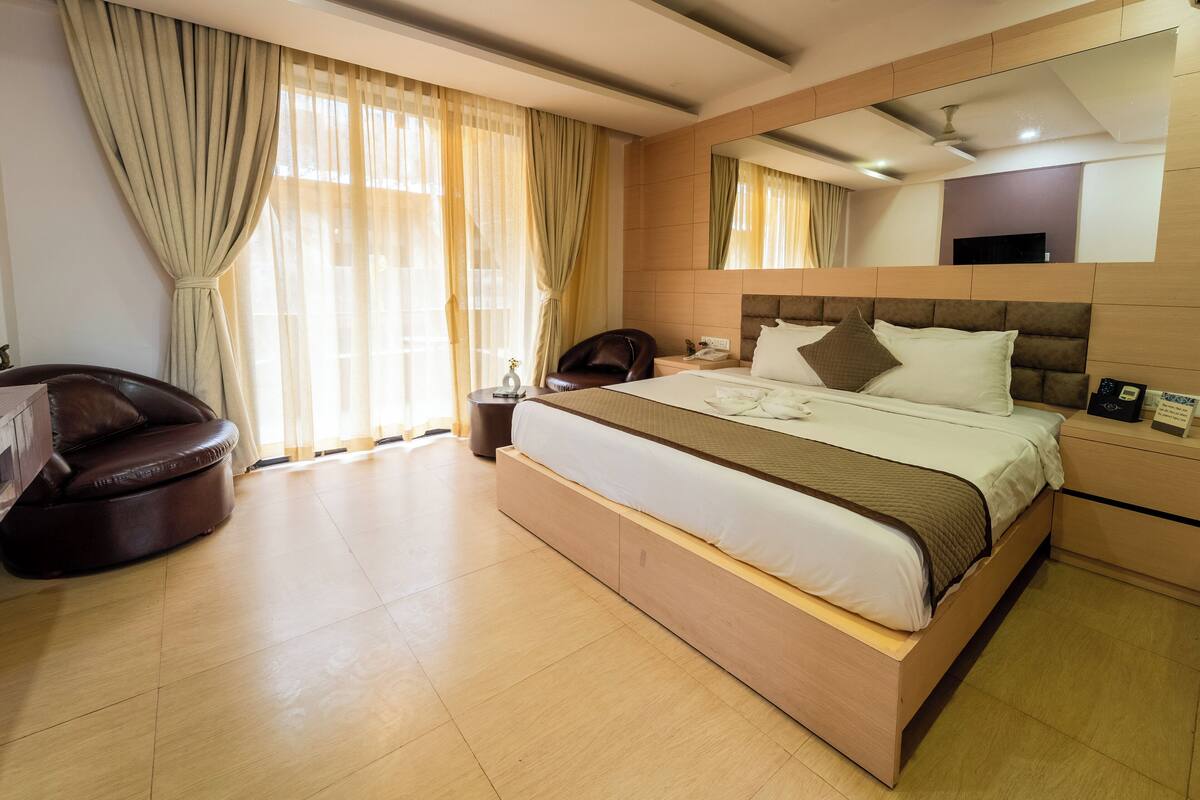 Una habitación de hotel moderna con una cama grande, un sillón de cuero, una mesita de noche con lámpara y un espejo grande en la pared.