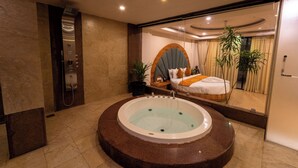 Un baño moderno con una bañera grande, ducha y un espejo que refleja un dormitorio con cama y cortinas.