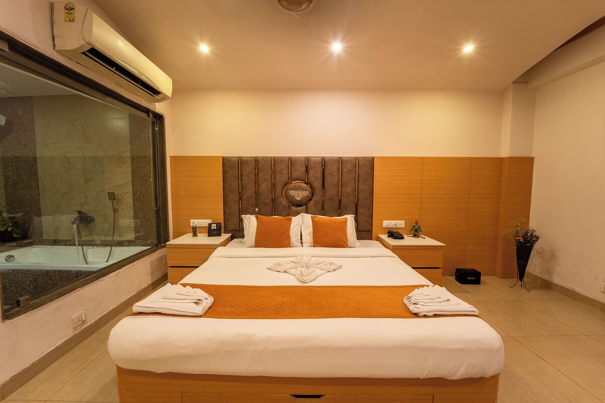 A hotel room with a large bed, a glass shower enclosure, wooden headboard, and a bedside table with a lamp.