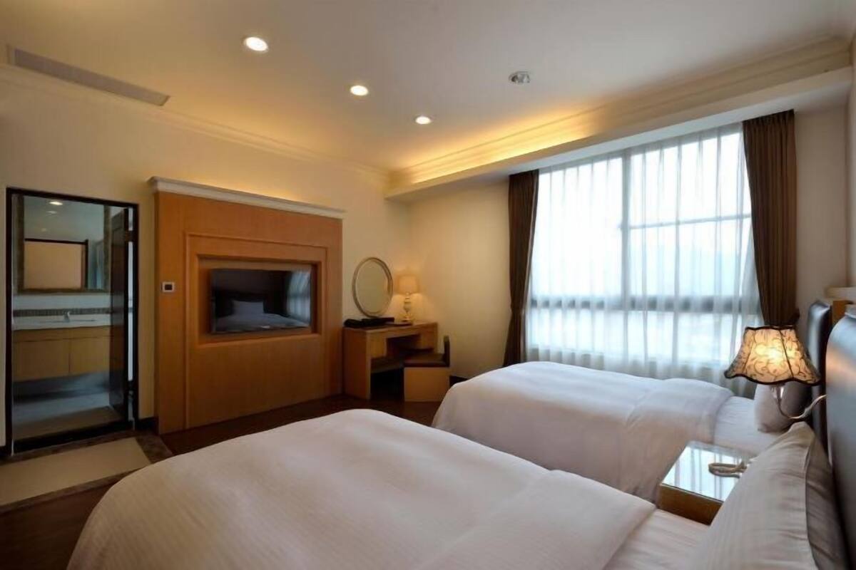 Une chambre d’hôtel avec deux lits, une télévision, un bureau avec une lampe, un miroir et une fenêtre avec des rideaux.