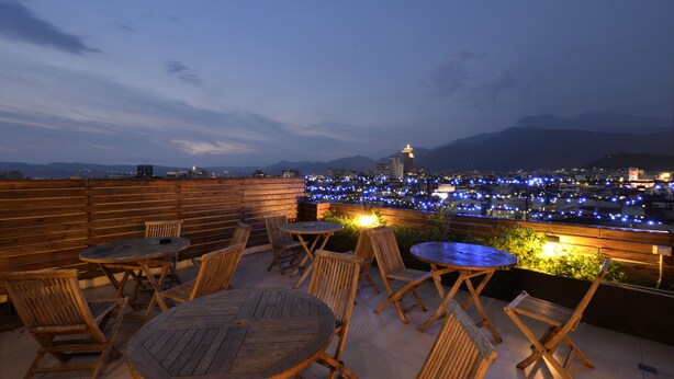A rooftop terrace with wooden furniture and a view of a cityscape at dusk.