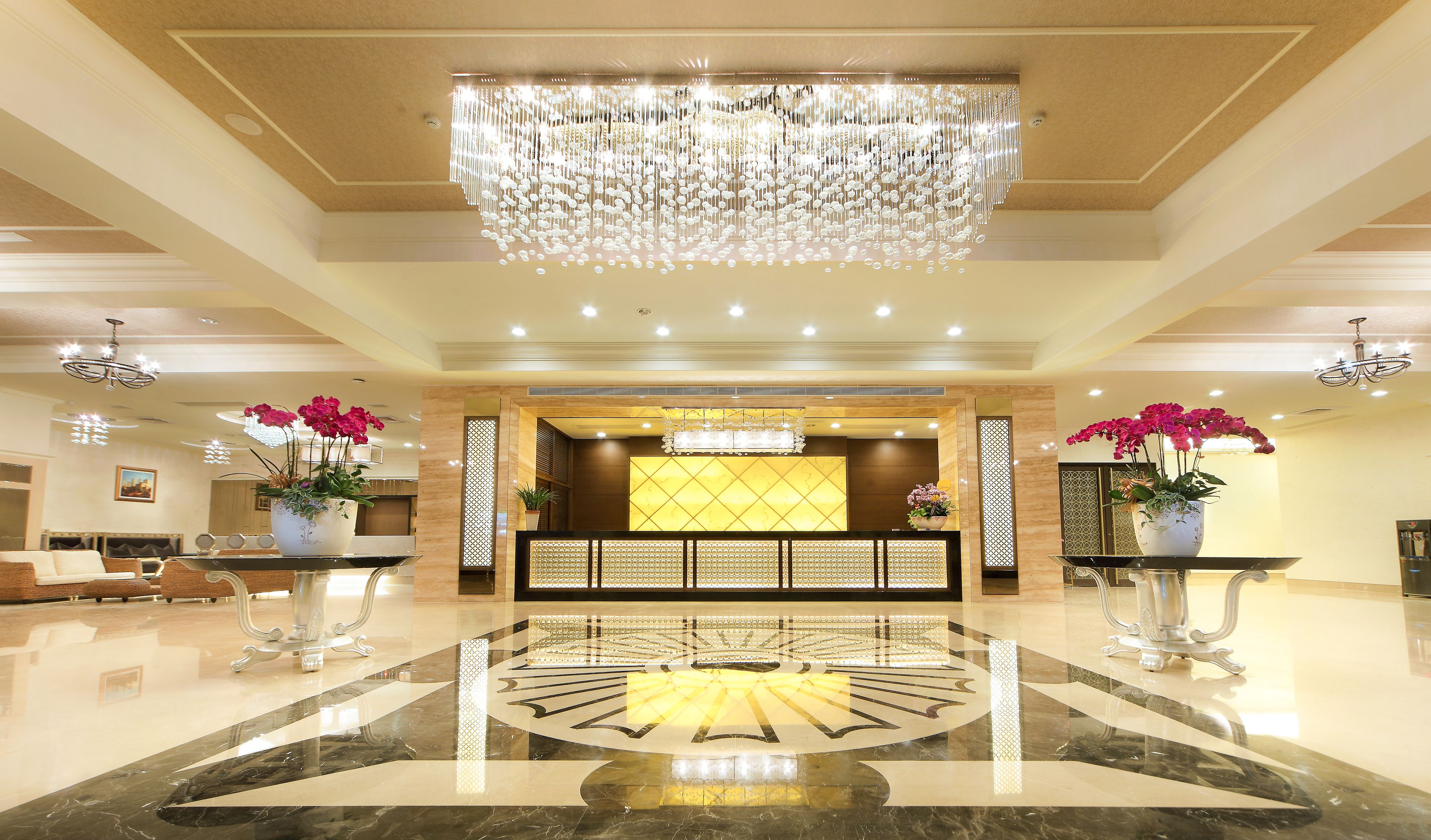A spacious lobby with a large chandelier, a reception desk, and potted orchids.