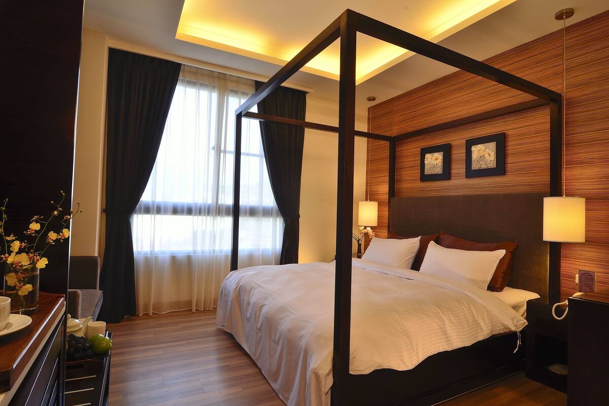 Un lit à baldaquin dans une chambre d’hôtel, avec une tête de lit en bois, une table de chevet avec une lampe et une fenêtre avec des rideaux.