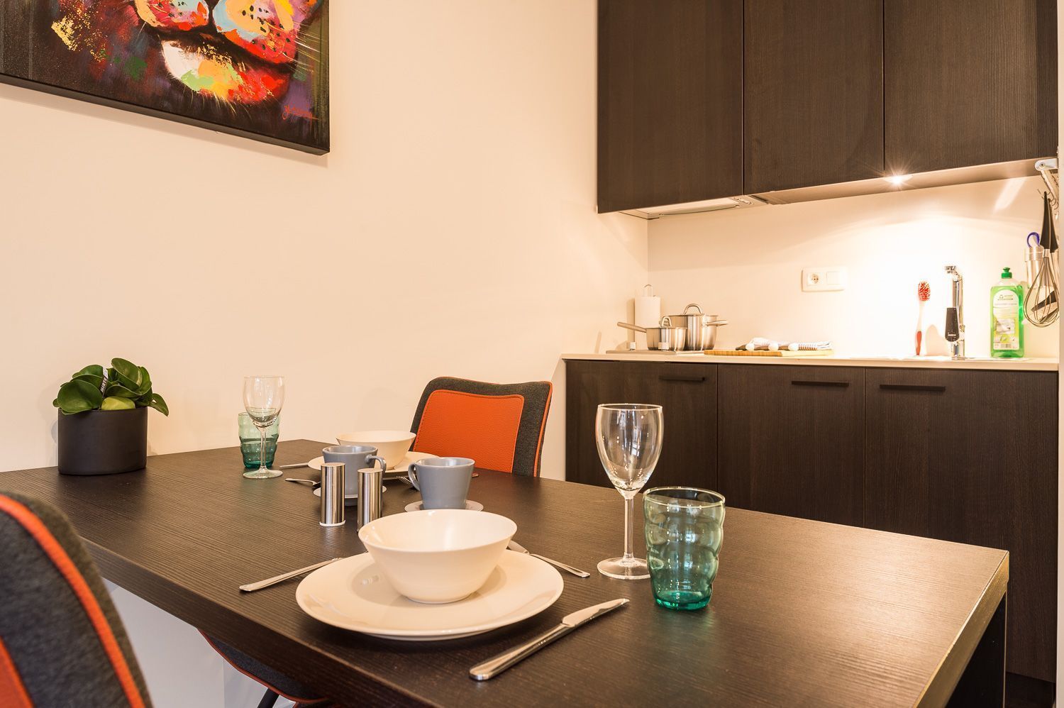 Un coin repas avec une table dressée pour deux personnes, une plante en pot et un tableau accroché au mur.