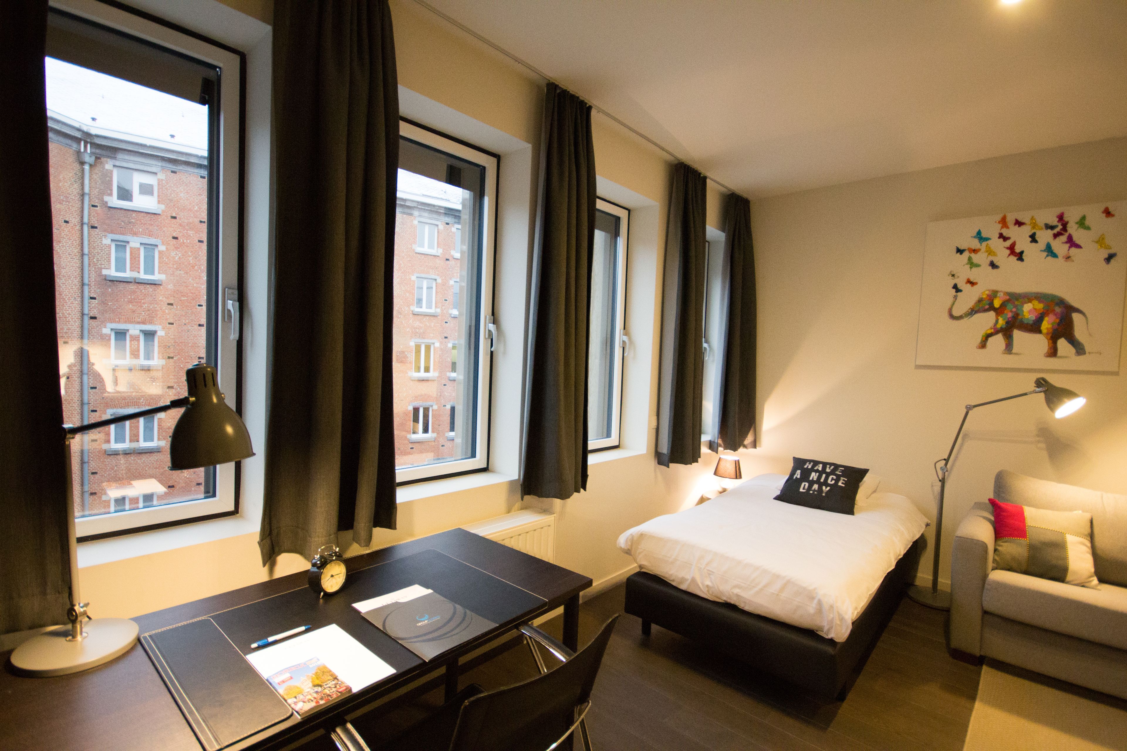 Una camera d'albergo con un letto, una scrivania, una sedia, una lampada e una finestra con tende.