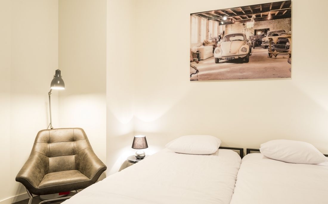 A bedroom with a bed, two pillows, a chair, a desk with a lamp, and a wall-mounted picture of vintage cars.