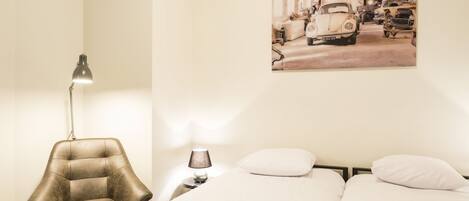 A bedroom with a bed, two pillows, a chair, a desk with a lamp, and a wall-mounted picture of vintage cars.