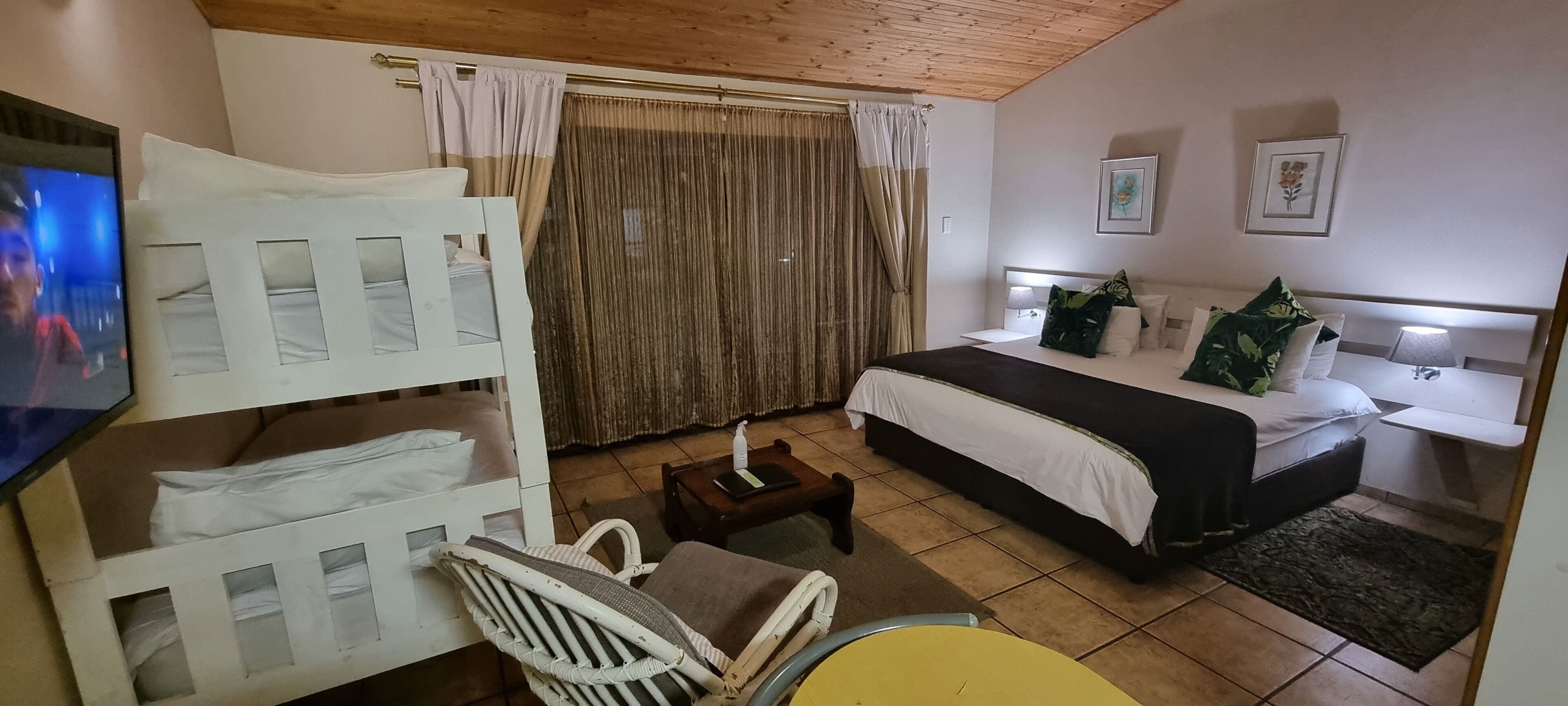 Quarto de hotel com uma cama beliche, uma cama de solteiro, uma mesinha com luminária, uma cadeira e uma TV fixada na parede.