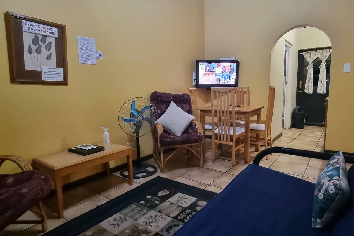 A compact living room with a sofa, a dining table with chairs, a TV, a fan, and a small table with a bottle and papers.