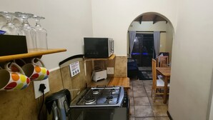 A compact kitchen with a gas stove, a microwave, and a small refrigerator. There are colorful mugs on a shelf and a dining table with chairs in the background.
