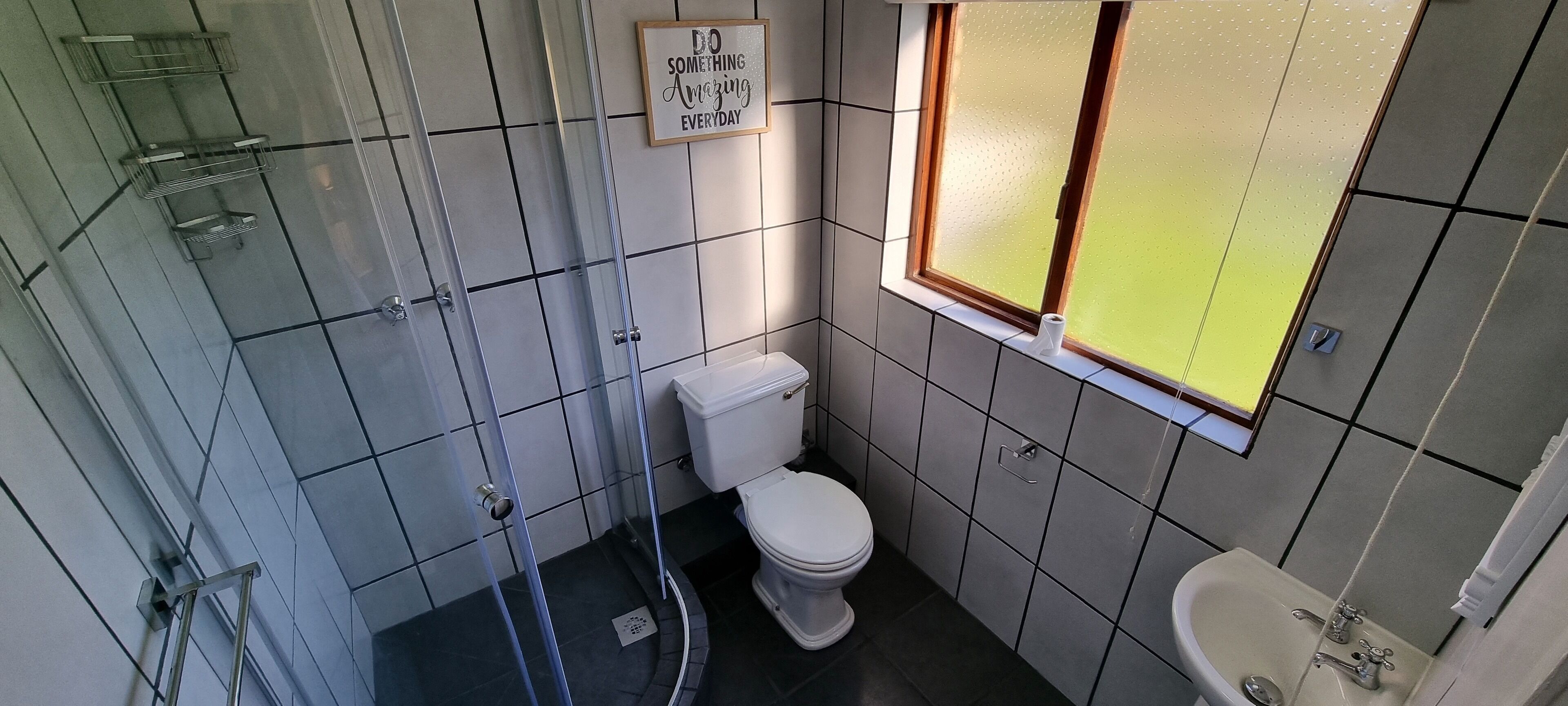 A bathroom with a toilet, sink, and shower. There is a motivational quote on the wall and a window with frosted glass.