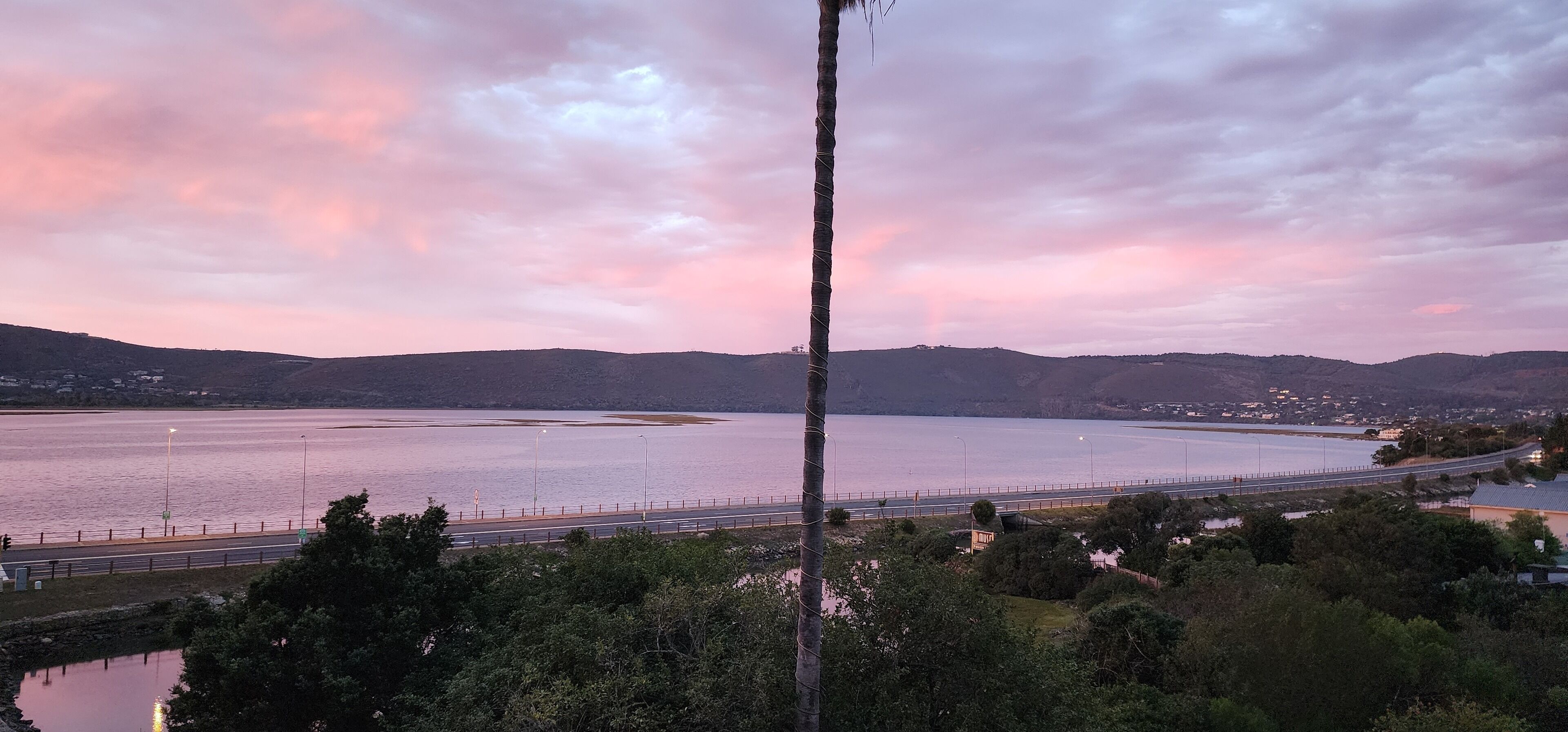 A coastal landscape with a road parallel to the water, surrounded by hills and greenery.