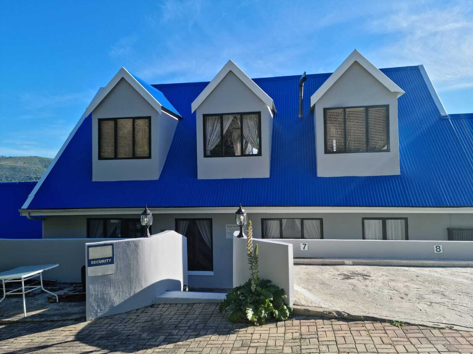 A two-story building with a blue roof and white walls, featuring three windows with shutters, a security sign, and a paved entrance area.