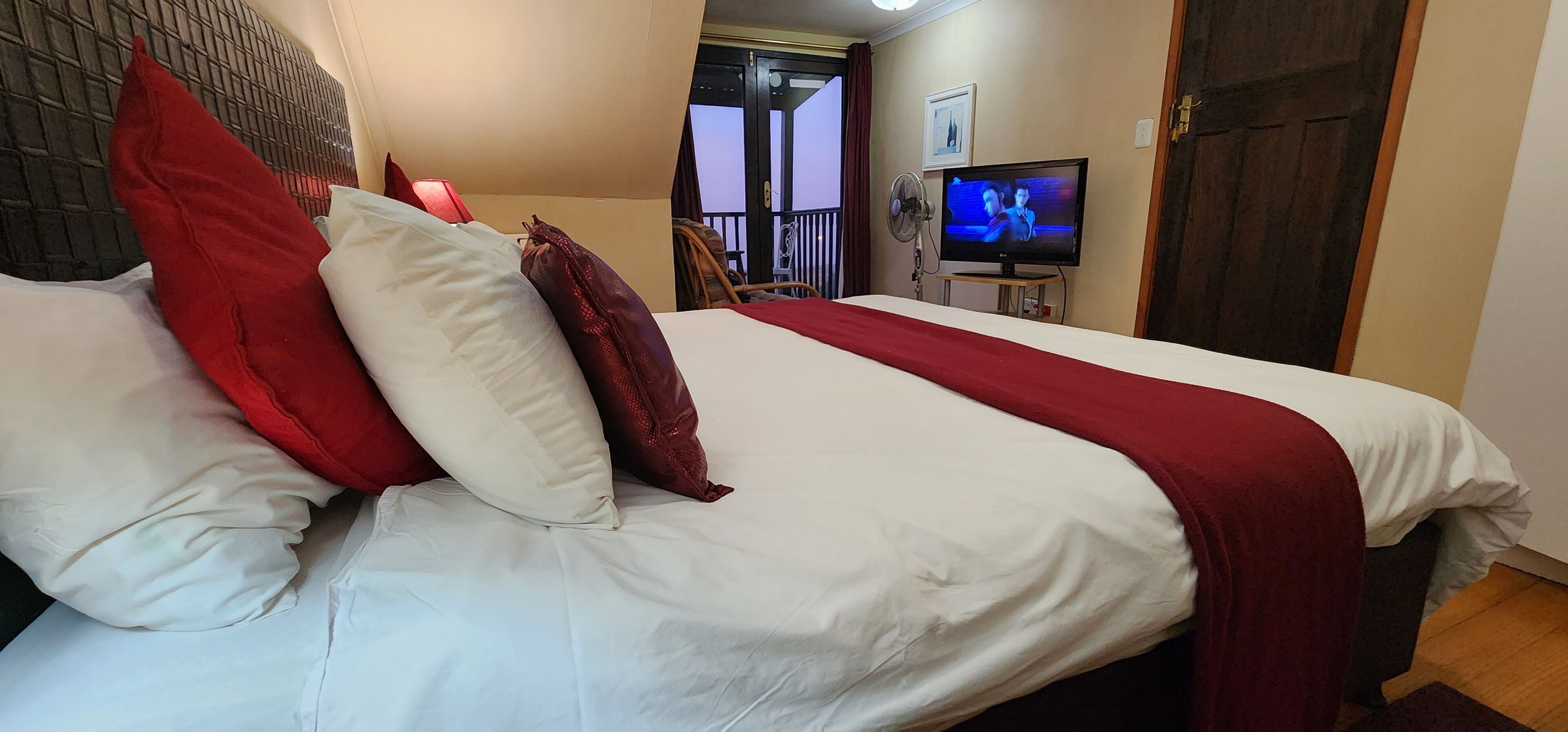 Una cama bien hecha con ropa de cama blanca y roja, un televisor sobre un mueble bajo y una puerta de madera al fondo.