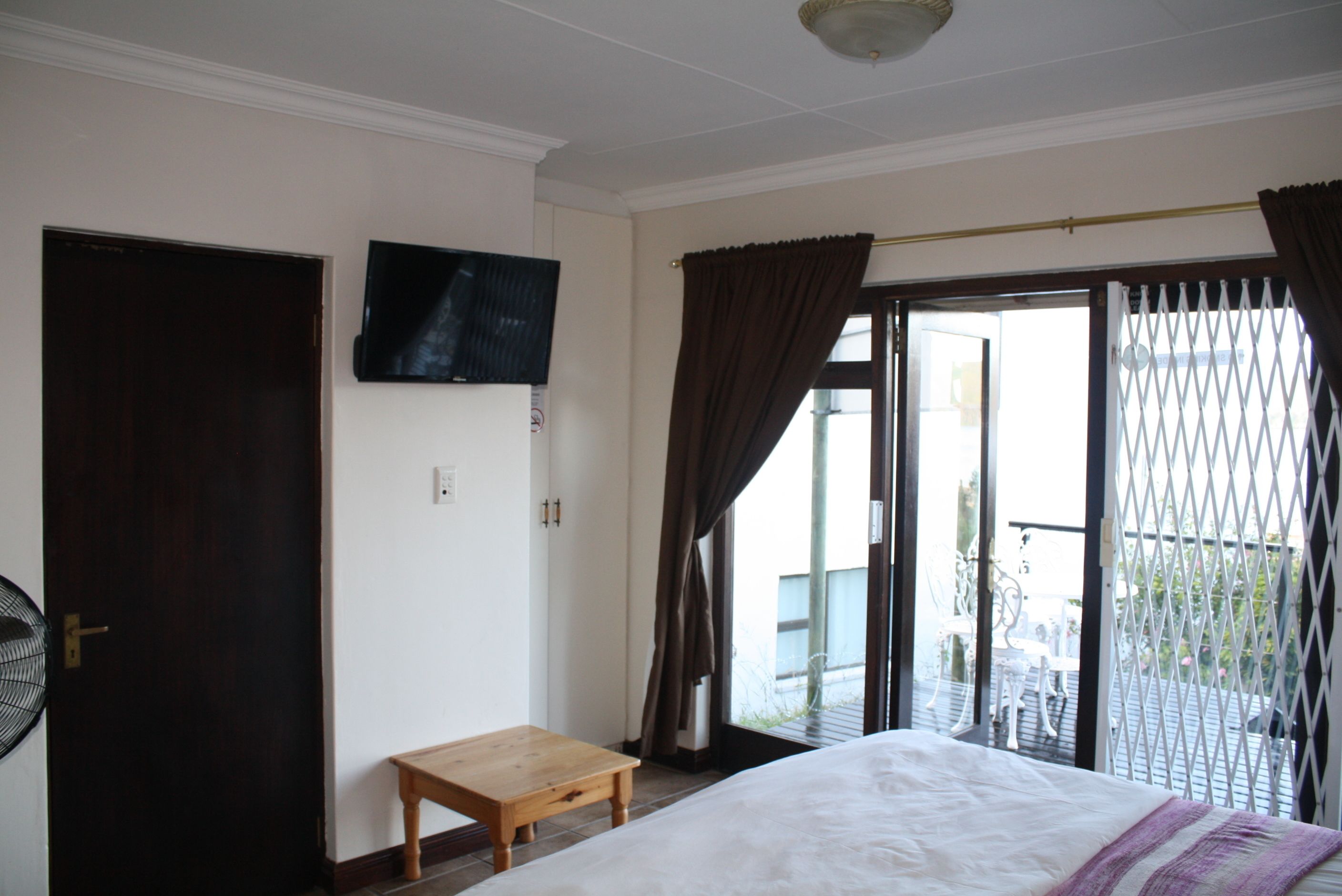 A bedroom with a bed, a wooden table, a TV mounted on the wall, and a sliding glass door leading to an outdoor area.