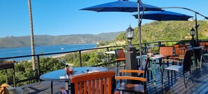 An outdoor dining area with tables and chairs, overlooking a body of water and hills. - Lagoon Lodge (Knysna)