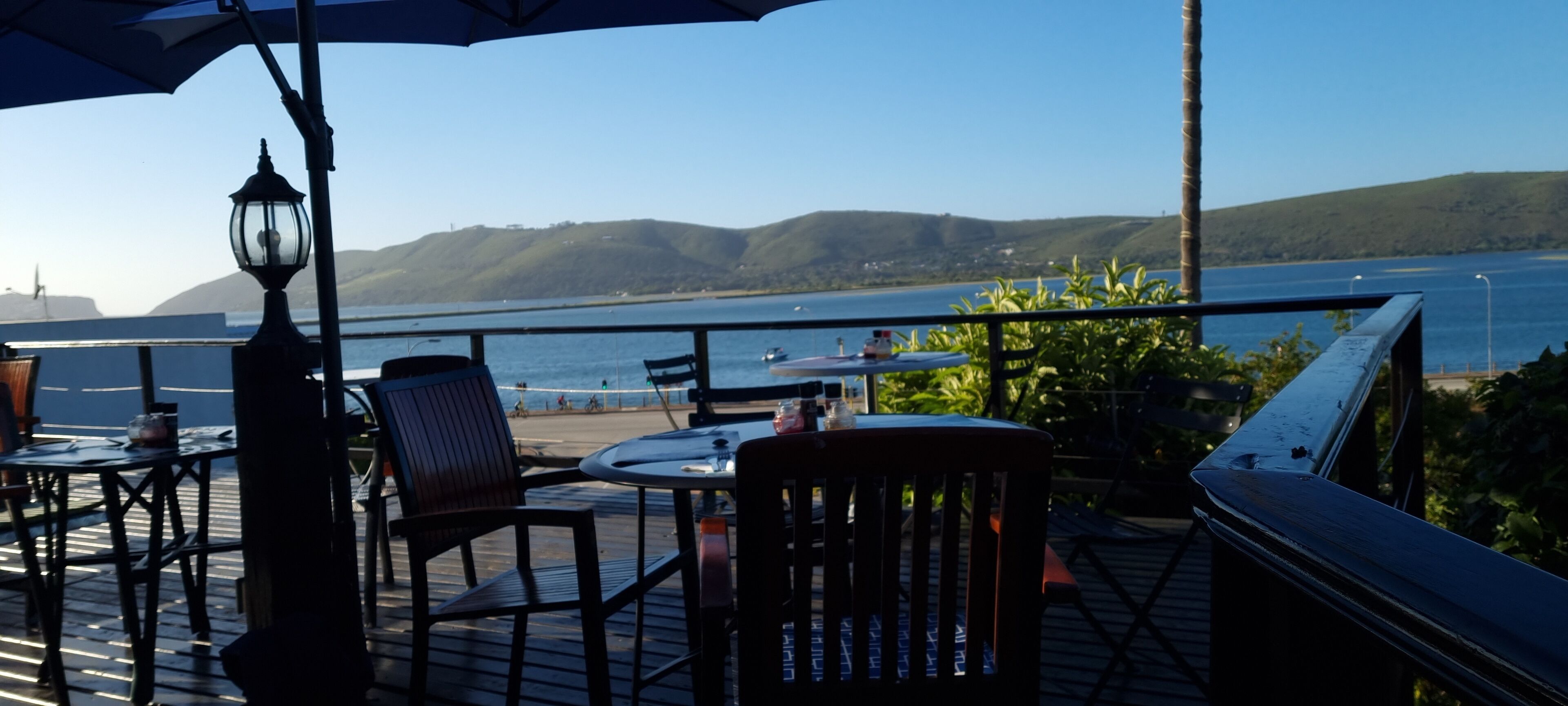 A deck with tables and chairs overlooking a body of water, with hills in the background.