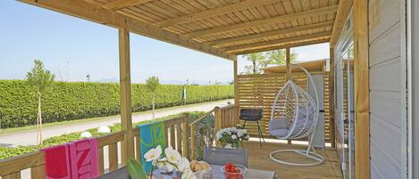 A covered outdoor dining area with a wooden deck, a table set for two, a hanging chair, and a view of a vineyard.